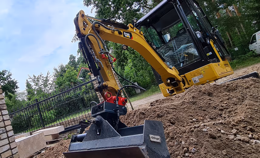 Gelber Caterpillar Minibagger mit Schaufel an einer Baustelle mit Erde und Bäumen im Hintergrund.