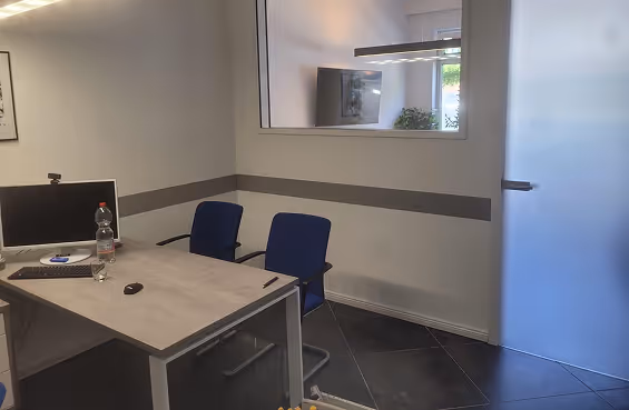 Ein modernes Büro mit einem Tisch, zwei blauen Stühlen, einem Computerbildschirm, einer Wasserflasche und einem Glas, mit einem Fenster im Hintergrund.