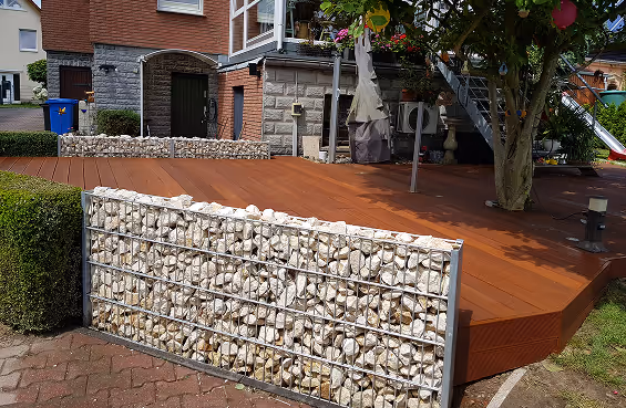 Garten mit modernem Holzdeck und Gabionen-Mauer aus Steinen vor einem Wohnhaus.