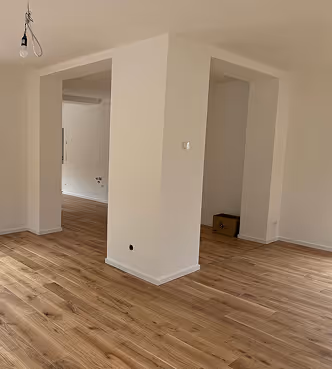 Leerer Raum mit weiß gestrichenen Wänden, Holzfußboden und einer Glühbirnenfassung ohne Lampe an der Decke.