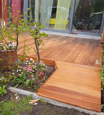 Holzterrasse vor einem Haus mit Glastüren, Blumenbeet und junger Pflanze im Garten.