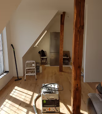 Helles Zimmer mit Holzboden, zwei Holzbalken, Fenster mit Sonnenlicht, Stuhl, Kühlschrank und Reinigungsgerät.