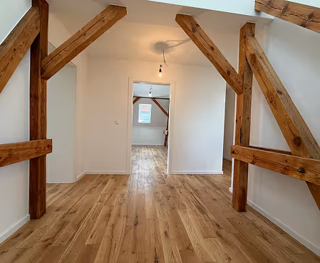 Leerer Raum mit hellem Holzboden, weißen Wänden und sichtbaren, massiven Holzbalken an den Wänden und der Decke.