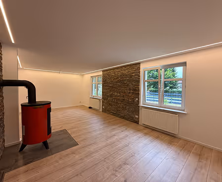 Leerer Raum mit hellem Holzboden, zwei Fenstern und rotem Kaminofen neben einer Wand mit Steinverkleidung.