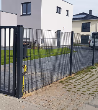 Schwarzer Metallzaun mit Tor vor modernem Haus mit Grüngras und gepflasterter Auffahrt.