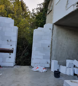 Baustelle mit unvollständiger weißer Mauer aus Betonsteinen und Baustoffen auf dem Boden, darunter Bäume im Hintergrund.