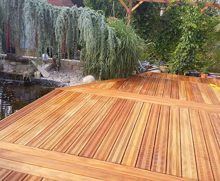Holzdeck mit verschiedenen Holztönen neben einem Gartenteich und grünen Pflanzen im Hintergrund.