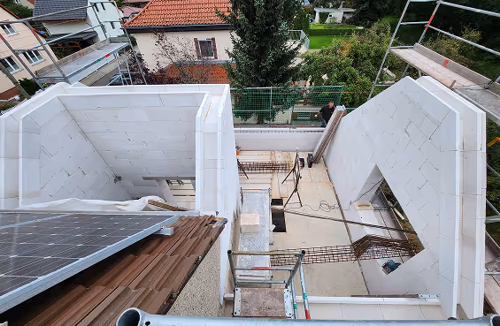 Bau einer Wohnstruktur mit weißen Ziegelwänden und Dächern mit Solarpanels, umgeben von Gerüsten und einem Garten im Hintergrund.