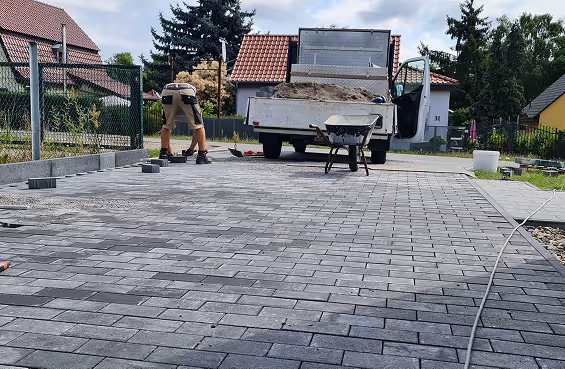 Mann verlegt graue Pflastersteine vor einem Lieferwagen mit Schubkarre auf gepflastertem Hof.