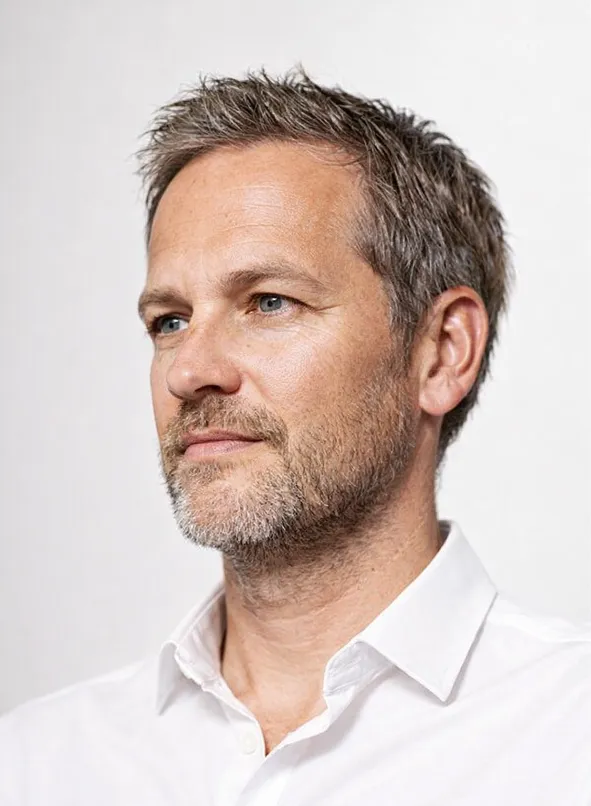 Middle-aged man with salt-and-pepper curly hair and beard wearing a dark crew-neck shirt looking slightly to the side.