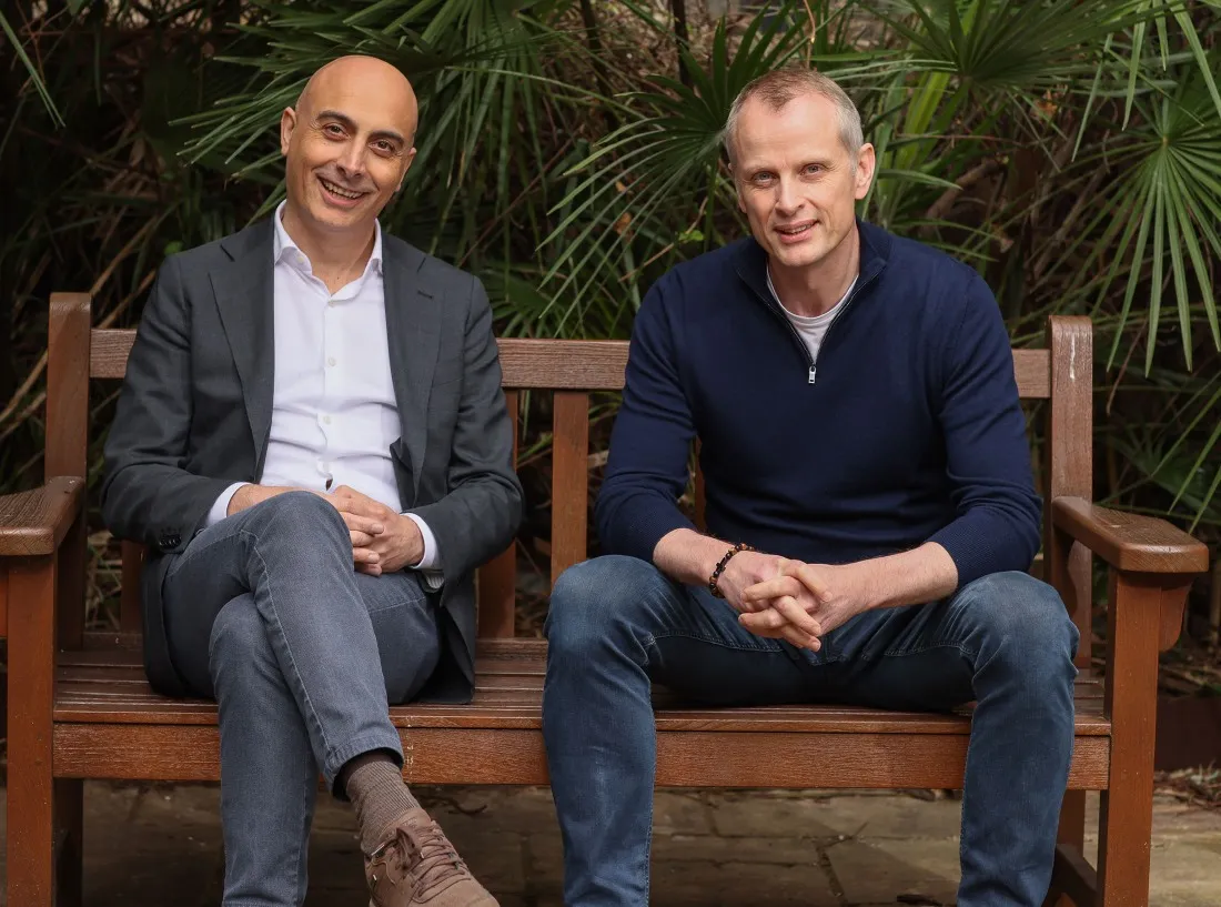 Two men sitting on a wooden bench outdoors with green foliage behind them, smiling at camera.