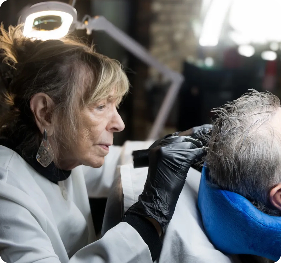 Woman wearing black gloves carefully working on a man's hair or scalp under a bright ring light.