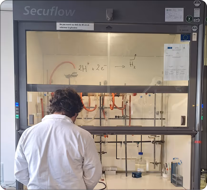 Chercheur de dos en blouse blanche travaillant sous une hotte de laboratoire avec un schéma chimique sur la vitre.