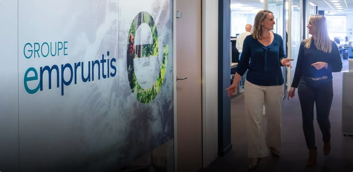 Deux femmes discutent en marchant dans un bureau près d'un mur avec le logo Groupe empruntis.