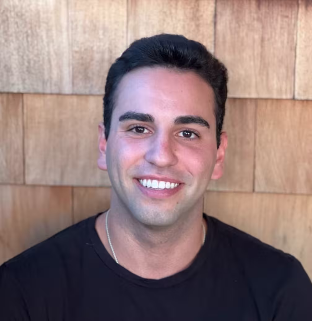 Smiling man with short dark hair wearing a black shirt against a wooden background.