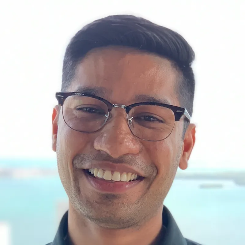Smiling man with glasses and short dark hair in front of a blurred blue and white background.