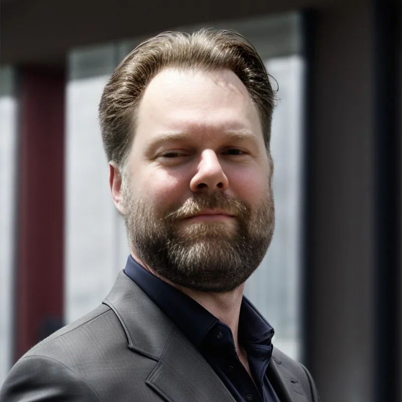 Bearded man in a dark suit jacket and shirt standing outdoors with a confident expression.