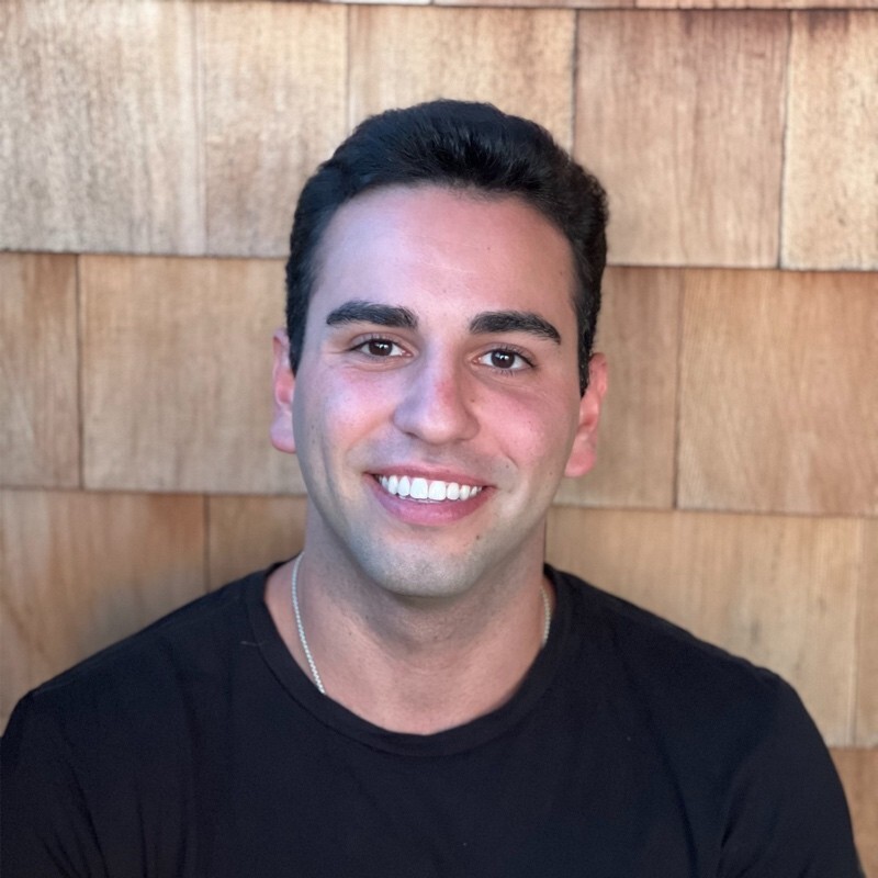 Smiling man with dark hair wearing a black shirt in front of wooden panel background.
