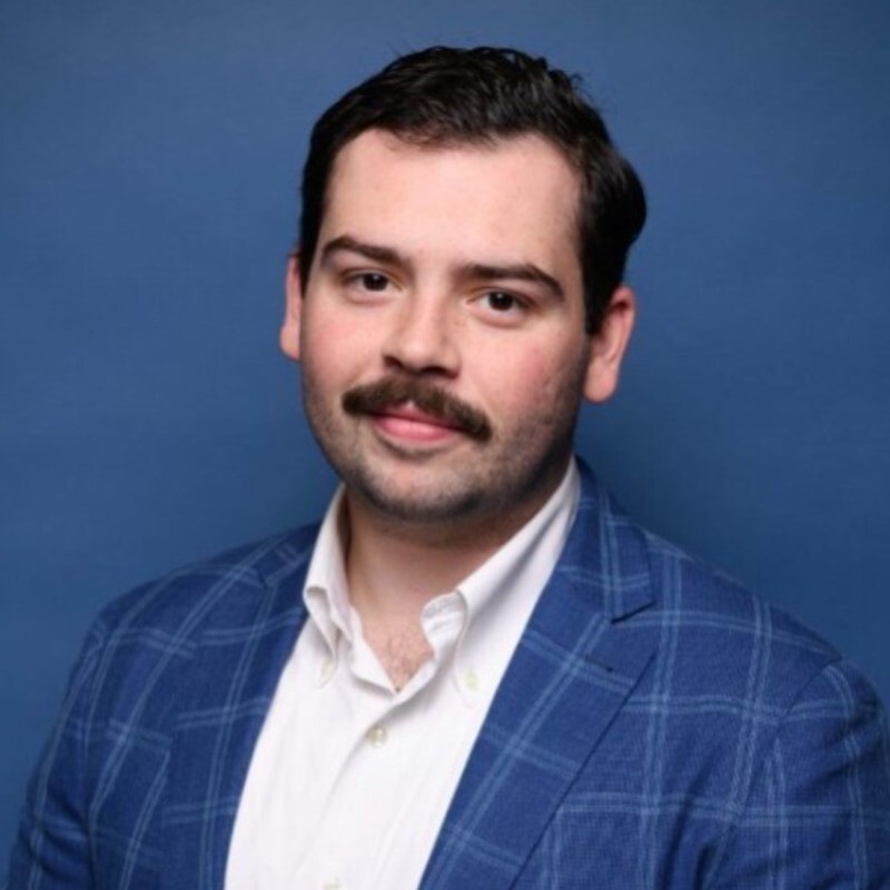 Man with dark hair and mustache wearing blue plaid blazer and white shirt against a blue background.