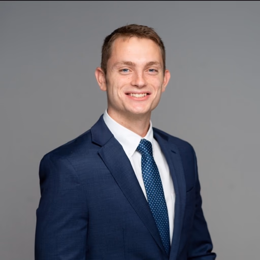 Young man in a navy suit, white shirt, and patterned tie smiling against a gray background.