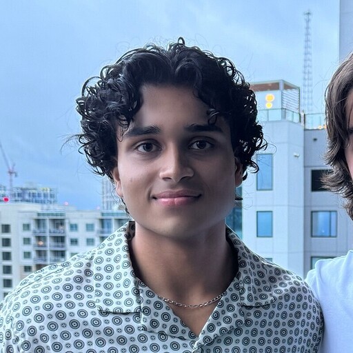 Young man with curly black hair wearing a patterned button-up shirt on a balcony in a city.