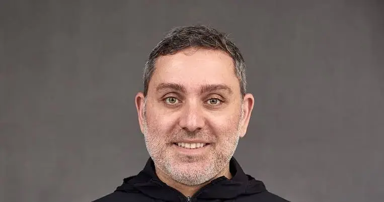 Smiling man with gray stubble, wearing a black zippered top, against a plain gray background.
