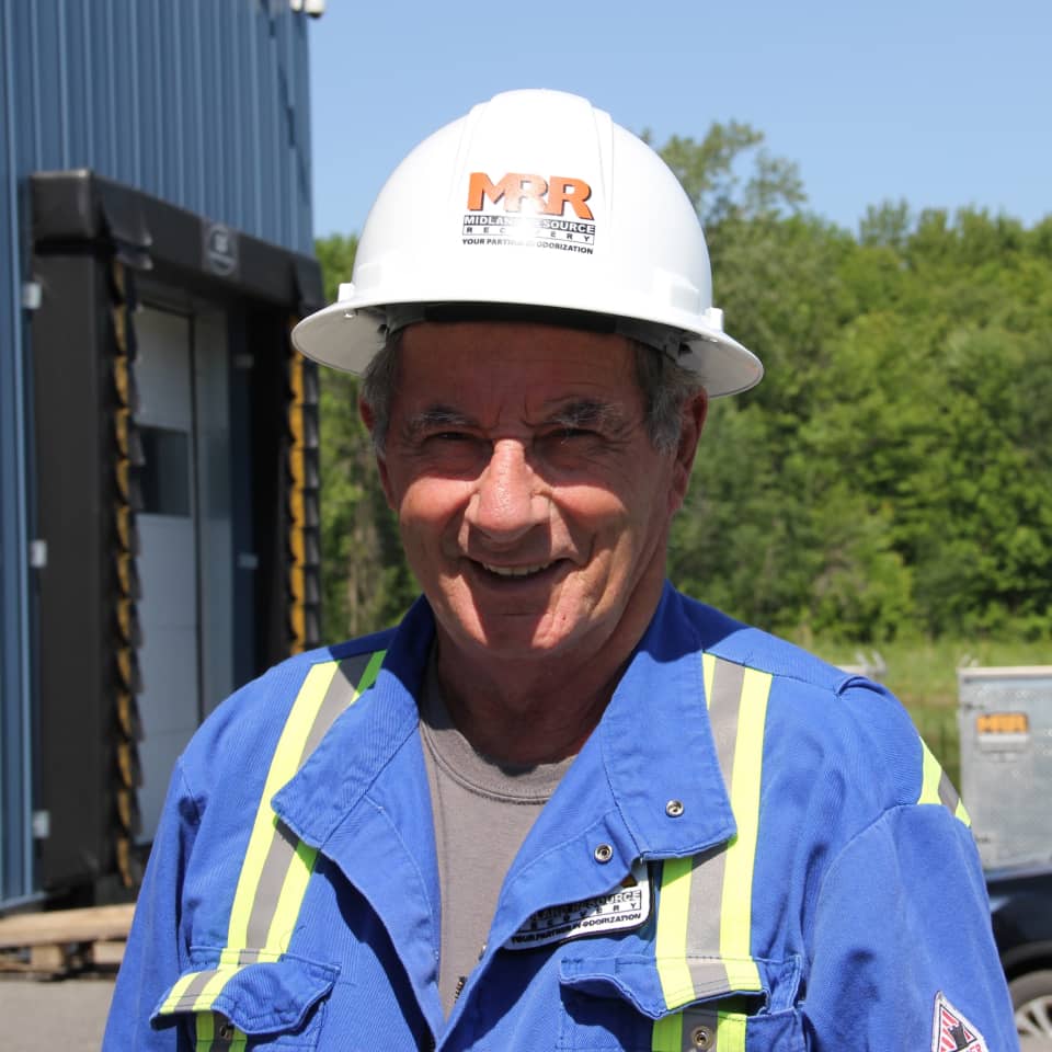 An MRR team member in a hardhat and safety jacket smiling in front of a company facility, from the timeline