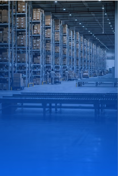 Large warehouse interior with tall metal shelving units filled with boxes, workers in safety gear, and conveyor belts for package handling under overhead lighting.