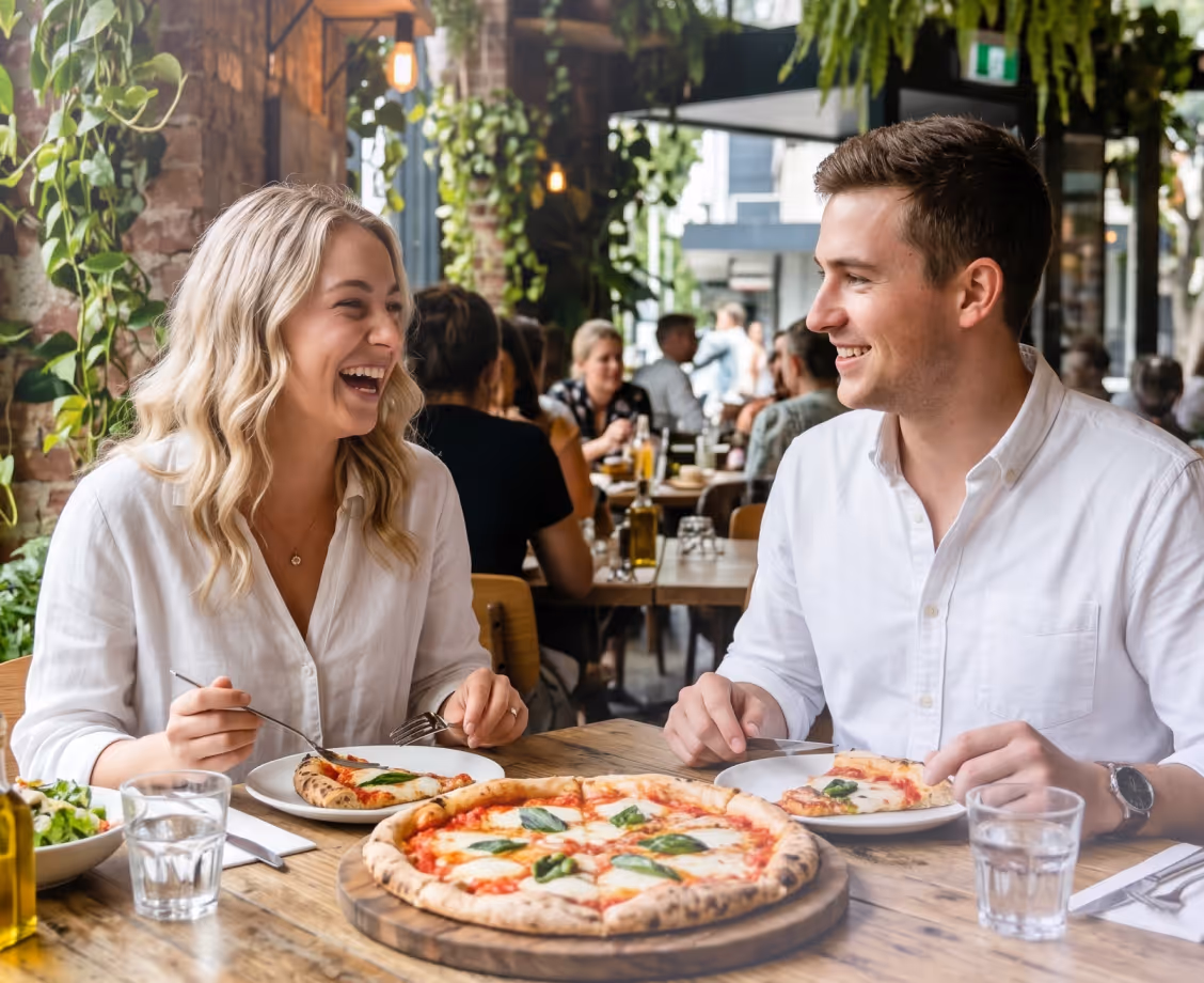 A smiling man and woman eating pizza at a wooden table in a lively restaurant with plants and other diners in the background.
