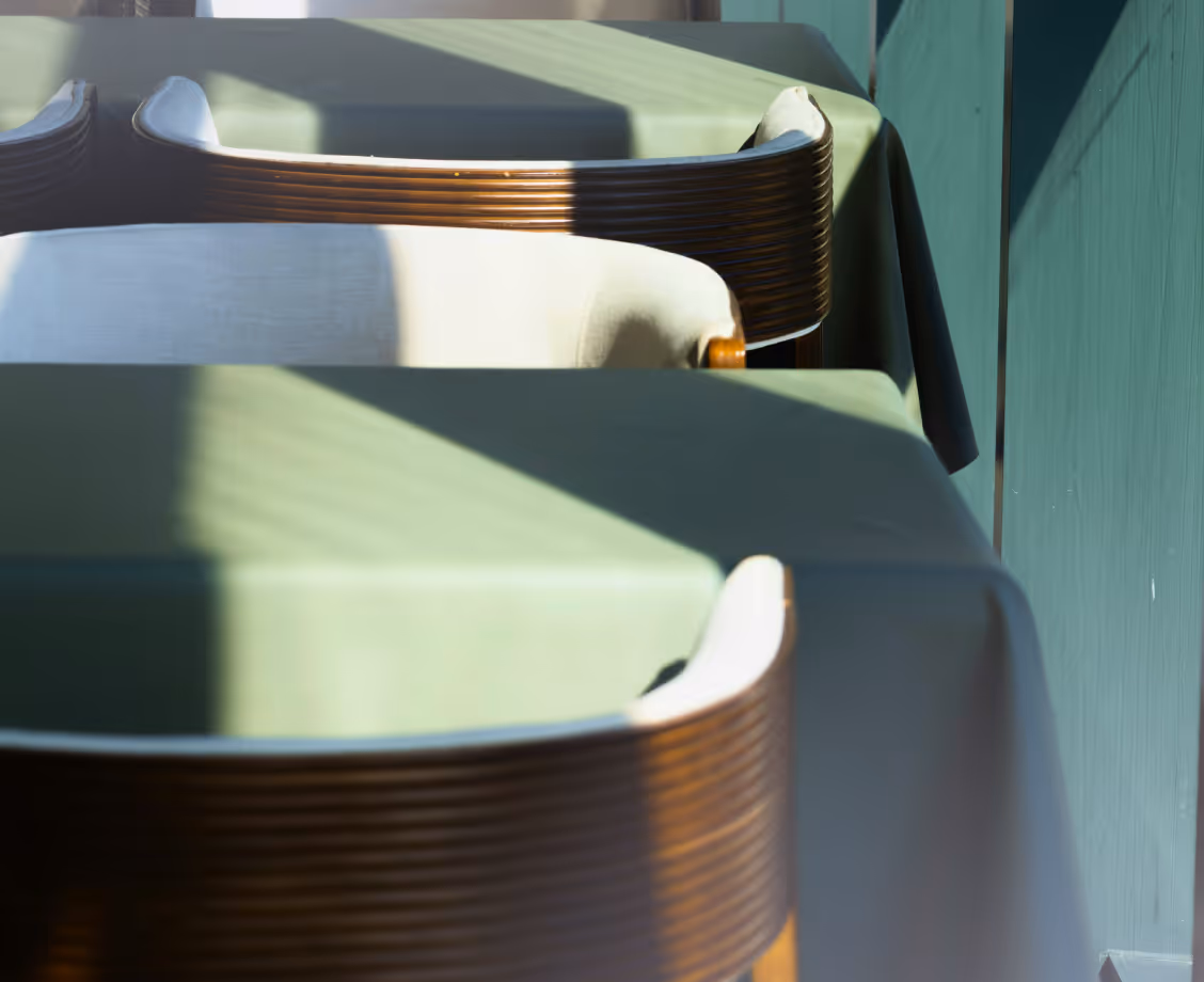 Sunlit green tablecloth-covered tables with wooden chairs lined up in a row.