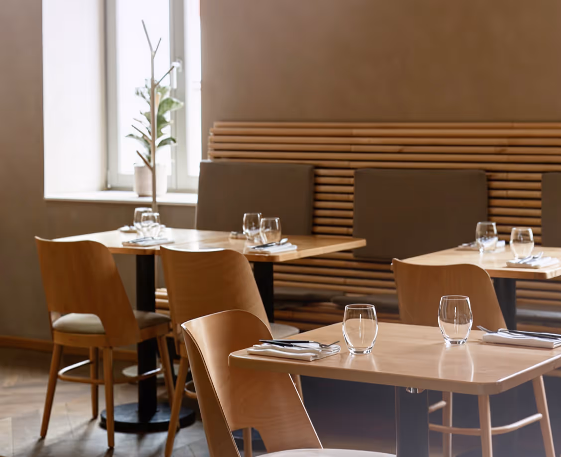 Modern restaurant interior with light wood tables and chairs, set with glasses, napkins, and cutlery.