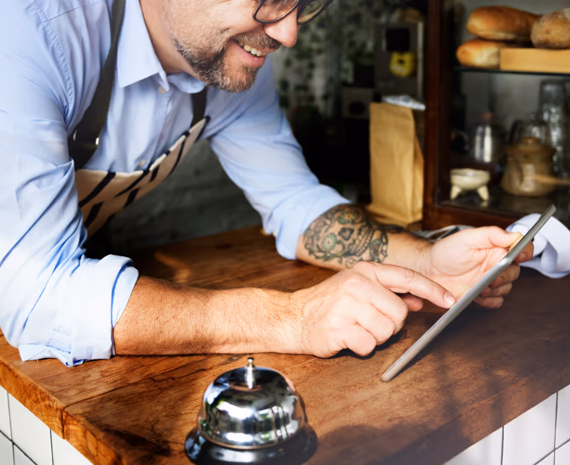 Smiling man in apron using tablet at wooden counter with service bell and bread in background.