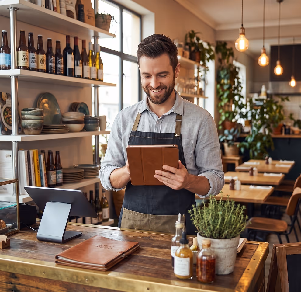Smiling barista wearing apron using tablet behind cafe counter with condiments and menus.