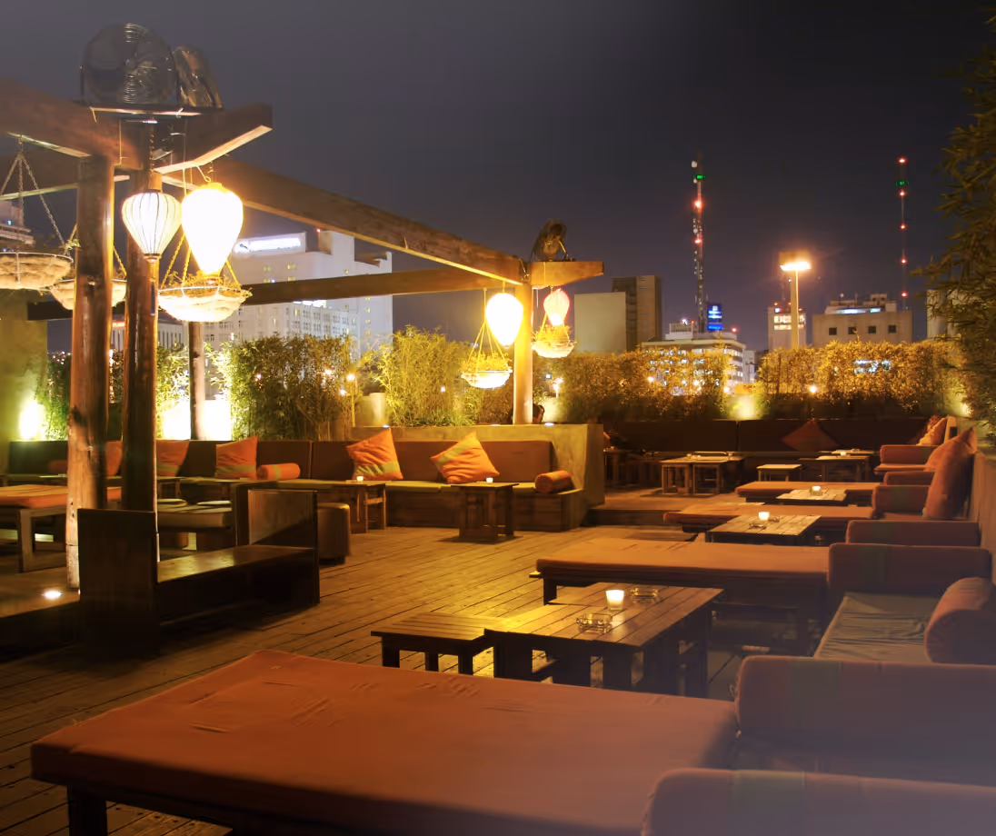 Cozy rooftop lounge with cushioned seating, hanging lanterns, and city lights in the background at night.