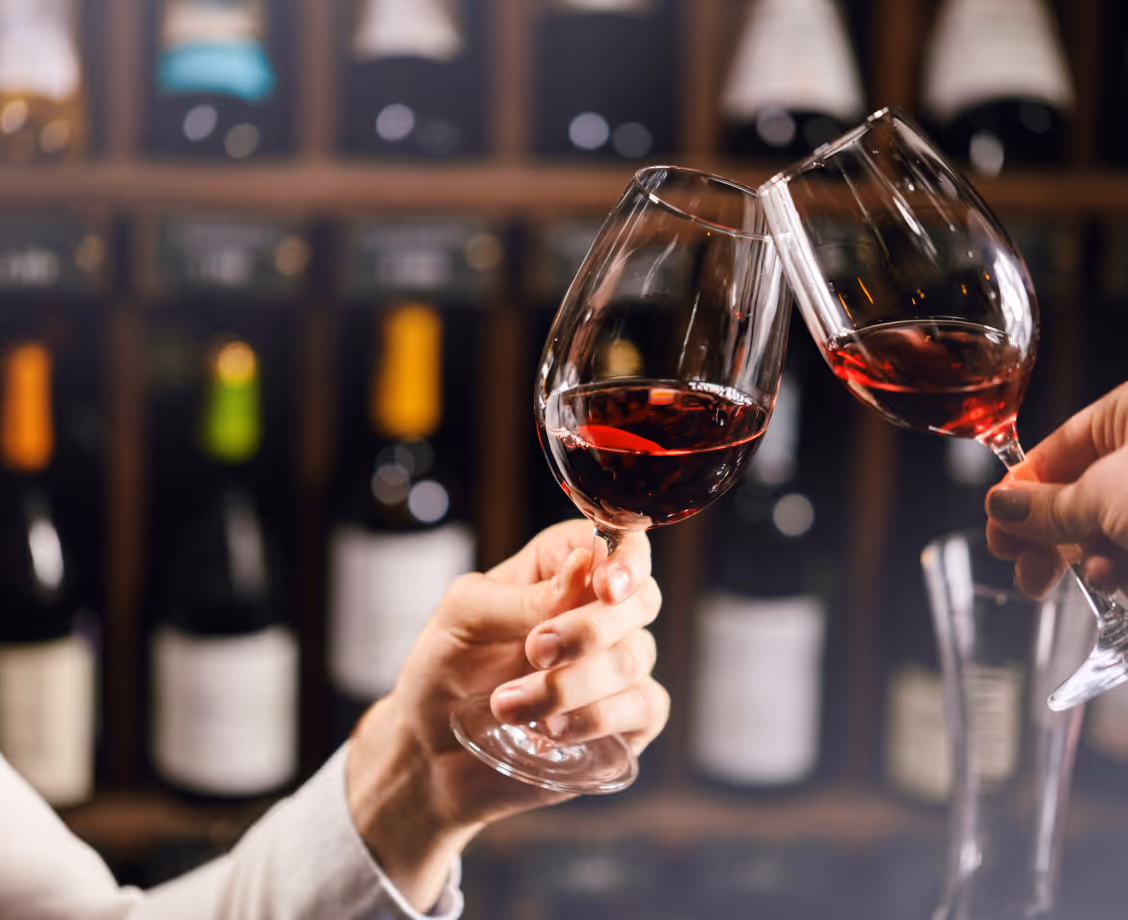 Two hands clinking glasses of red wine in front of a blurred wine rack background.