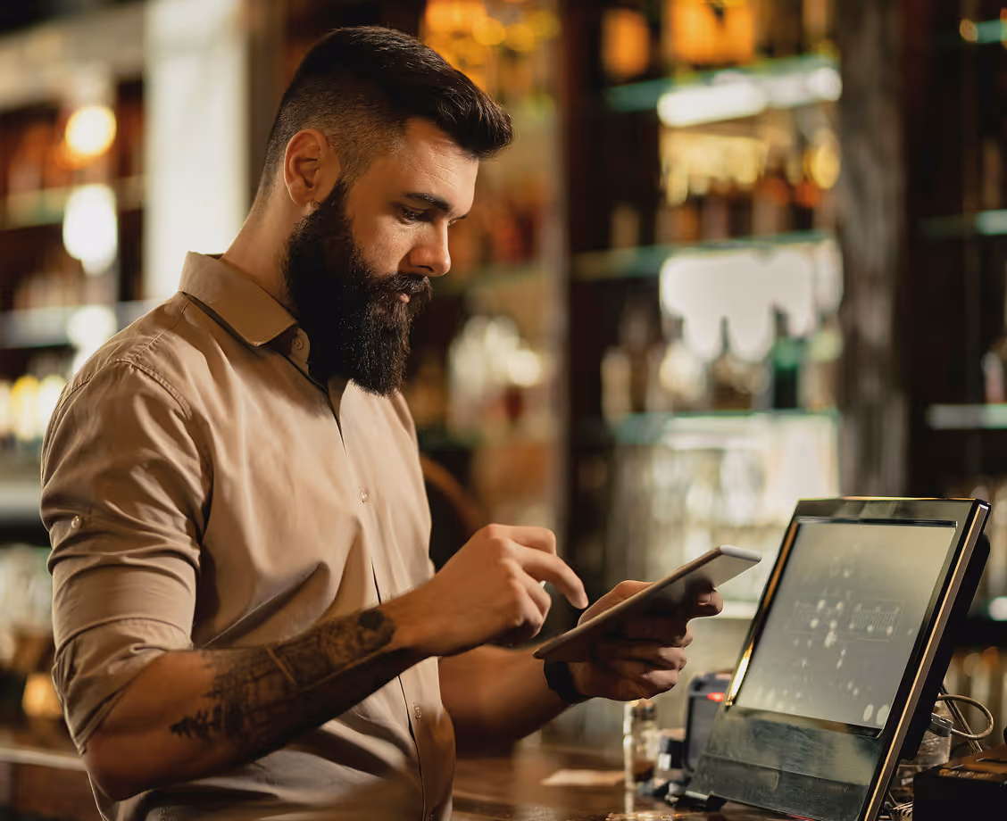 Man with beard using a digital tablet at a bar counter with a touchscreen register.
