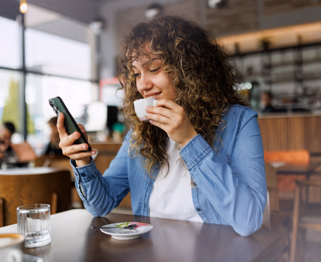 Curly-haired woman in blue shirt watches phone and sips coffee in a cozy cafe.