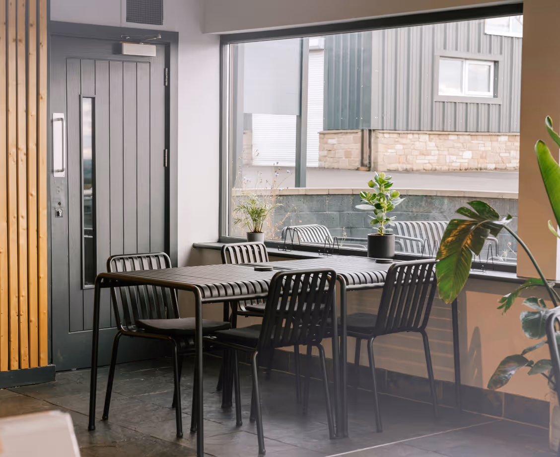 Modern dining area with black metal table and chairs near a large window showing outdoor industrial building.