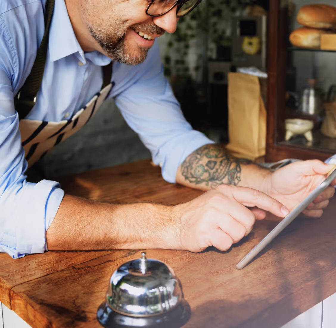 Man in apron with tattoo on arm using a tablet at a wooden counter near a service bell.