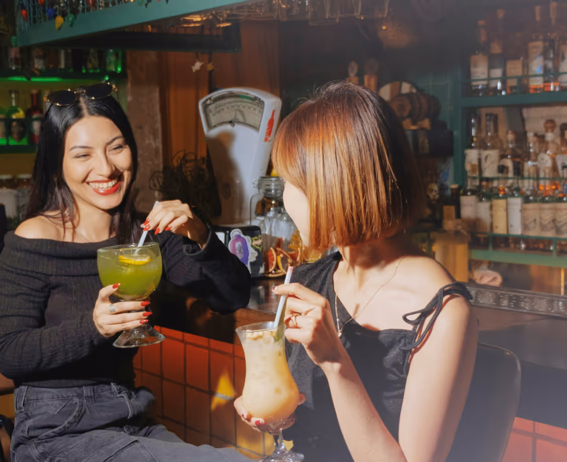 Two women enjoy colorful cocktails and smile while sitting at a bar with bottles in the background.