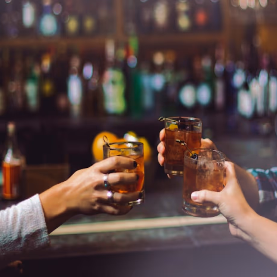 Three hands holding cocktails in short glasses, toasting at a bar with bottles blurred in background.