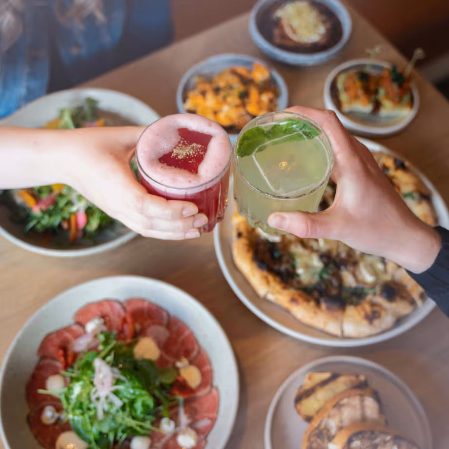 Two hands clinking cocktail glasses above a table with pizza, salad, and appetizers.