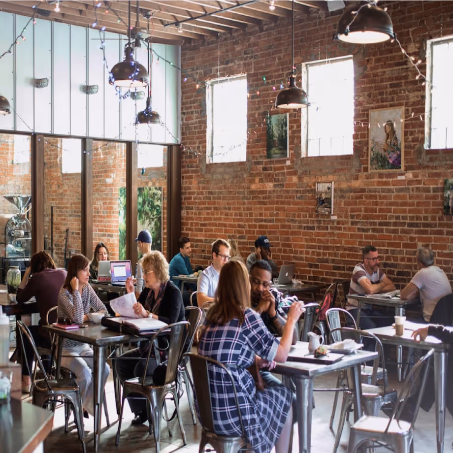 People sitting at tables chatting and working on laptops in a brick-walled café with hanging lights.