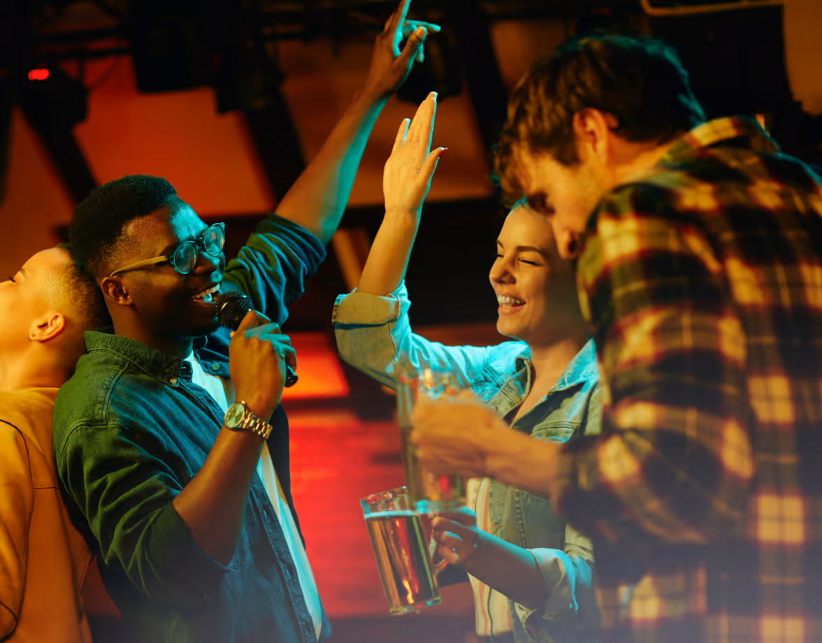 Group of friends enjoying karaoke in a bar with drinks and high-fiving joyfully.