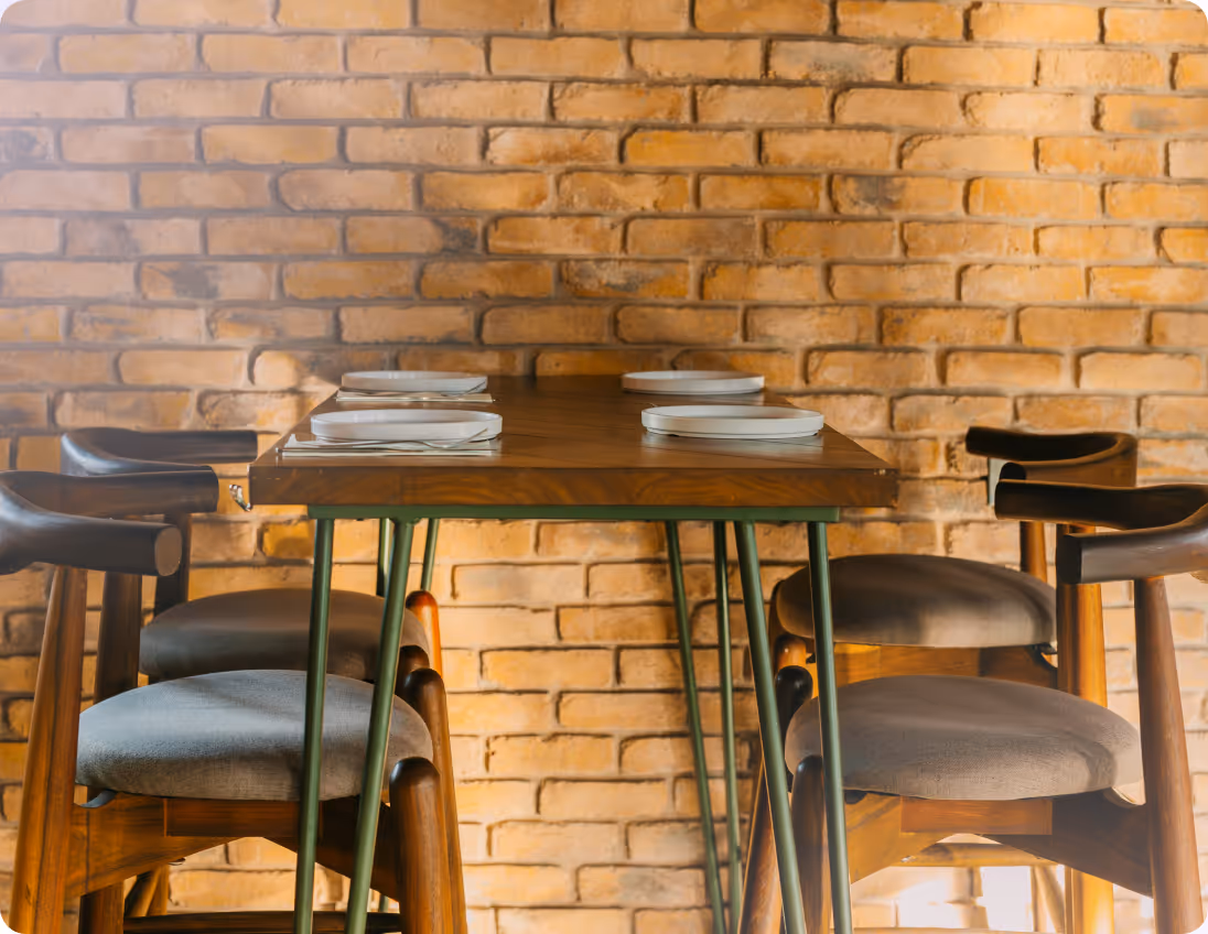 Wooden table set for four with white plates and gray cushioned chairs against brick wall.