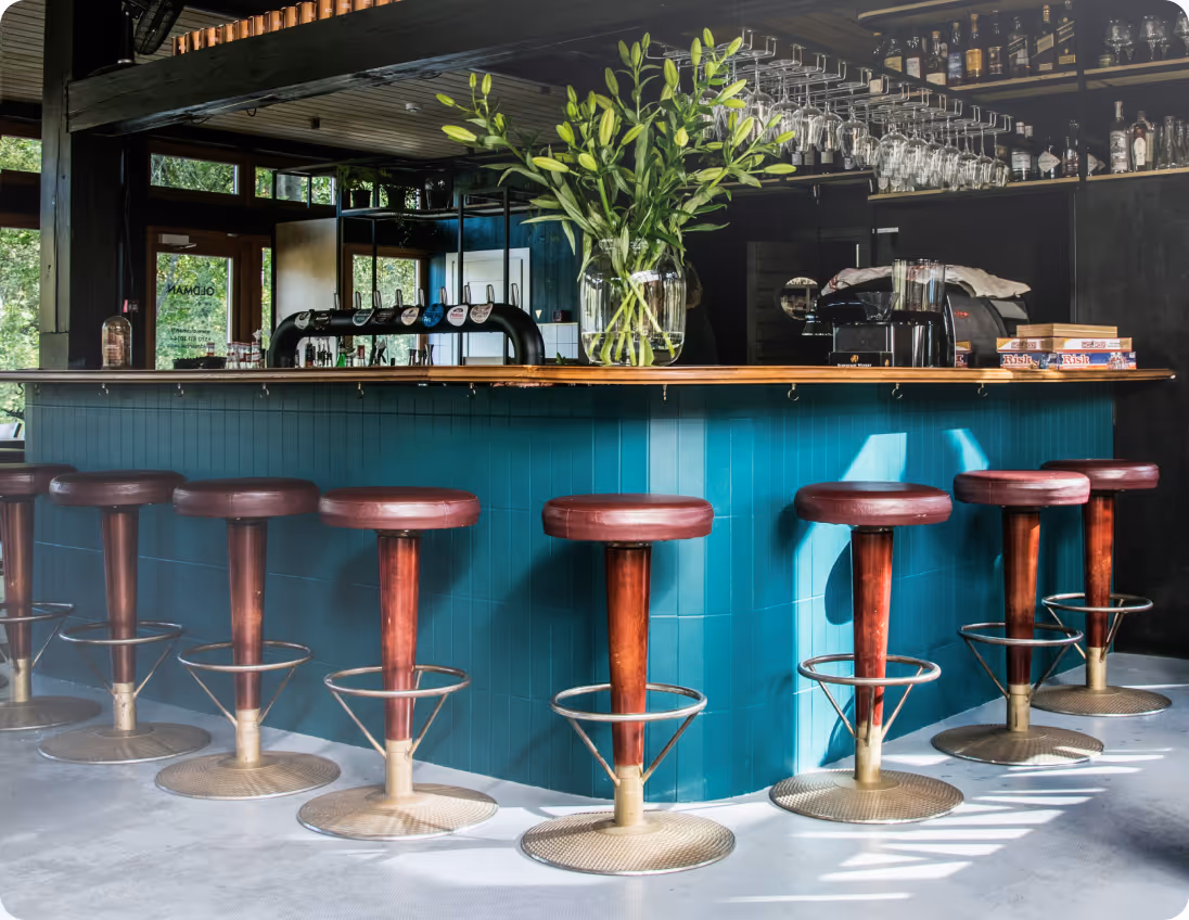 Modern bar with teal counter, red bar stools, hanging glasses, and a large green plant centerpiece.