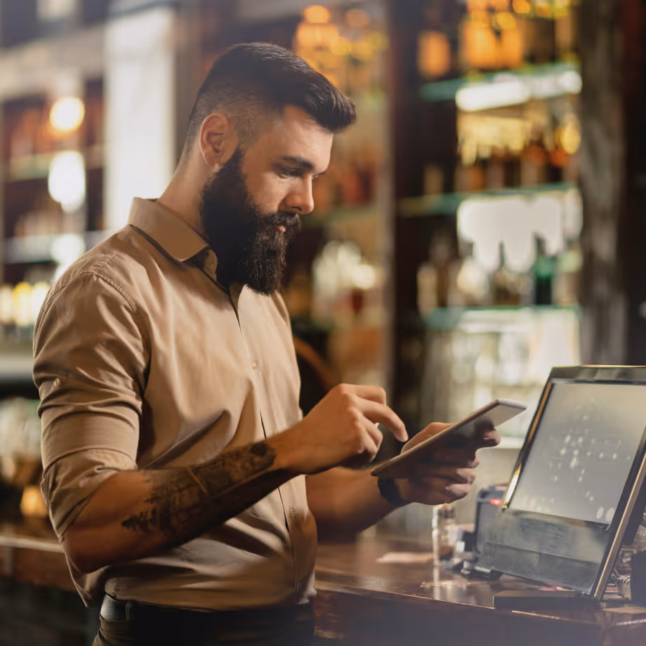 Bearded man using tablet at bar counter with a point of sale screen in the background.
