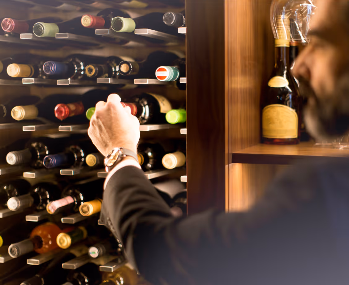 Man reaching for a bottle of wine in a wooden wine rack filled with various wine bottles.