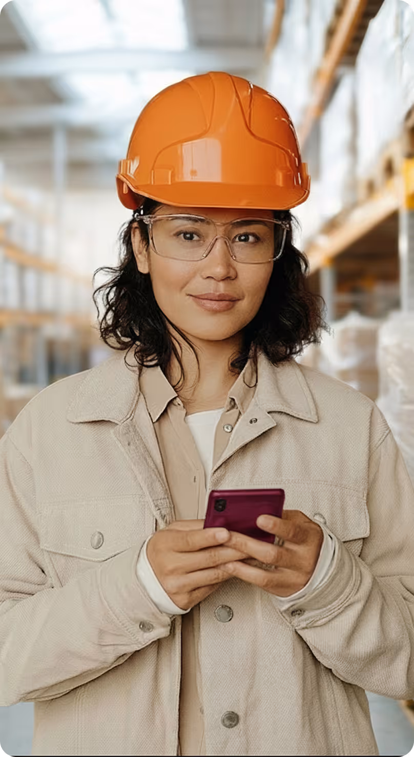 Frau in orangefarbenem Schutzhelm und Schutzbrille benutzt Smartphone in Lagerhalle.