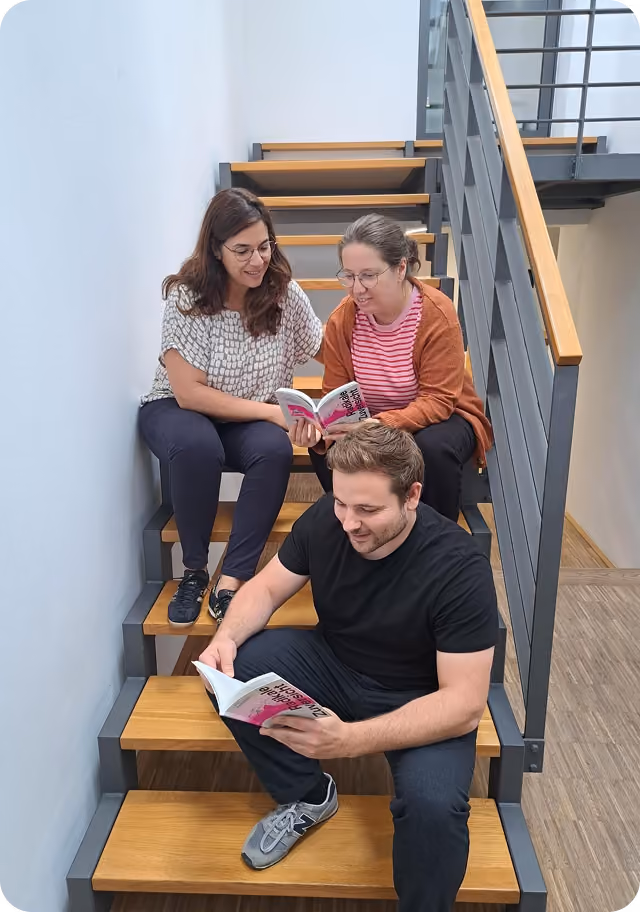 Drei Personen sitzen auf einer Treppe und lesen jeweils ein Buch gemeinsam oder allein.